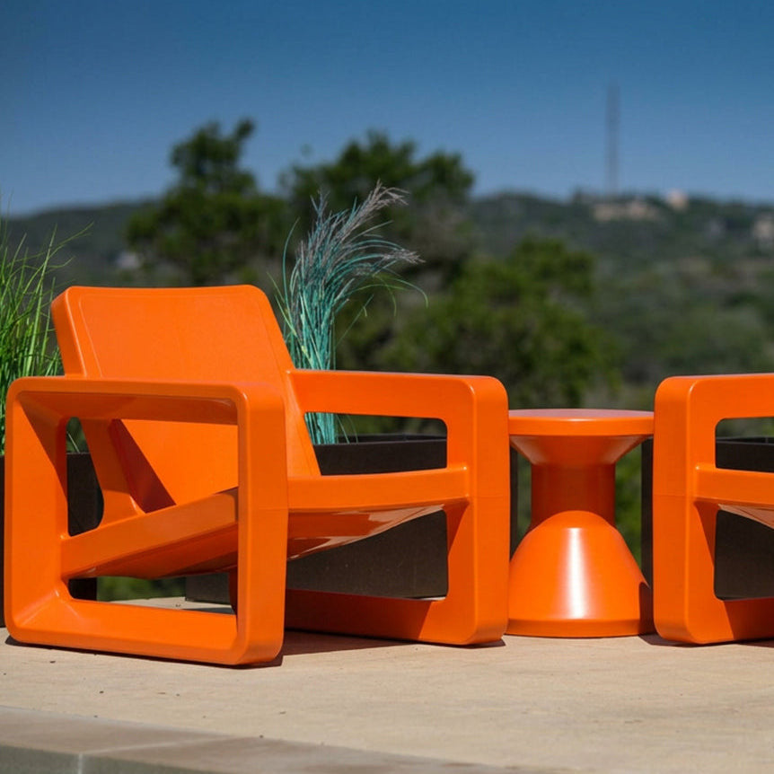 Bright orange outdoor patio furniture set including two modern armchairs and a small round side table, arranged on a stone patio with plants and a blurred mountain background.