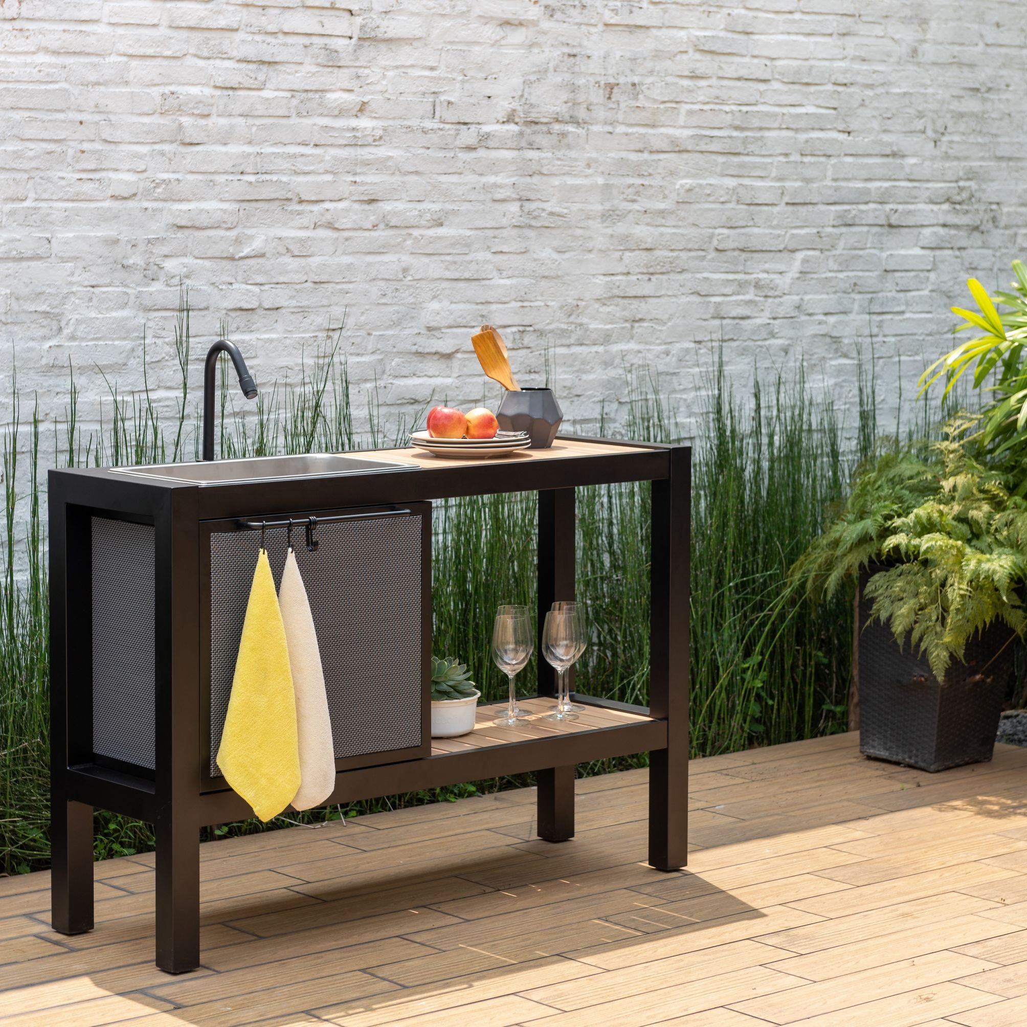 Outdoor kitchen island with sink, shelves, and plants against a stone wall.