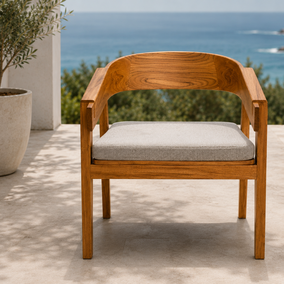 A wooden outdoor armchair with a curved backrest and a light gray textured seat cushion, positioned on a stone patio with a scenic ocean and rocky coastline in the background.