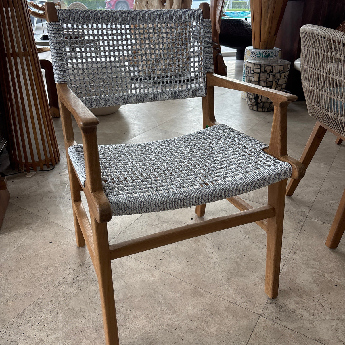 A wooden armchair with woven gray rope seating and backrest, photographed in a furniture showroom with other chairs and decor visible.