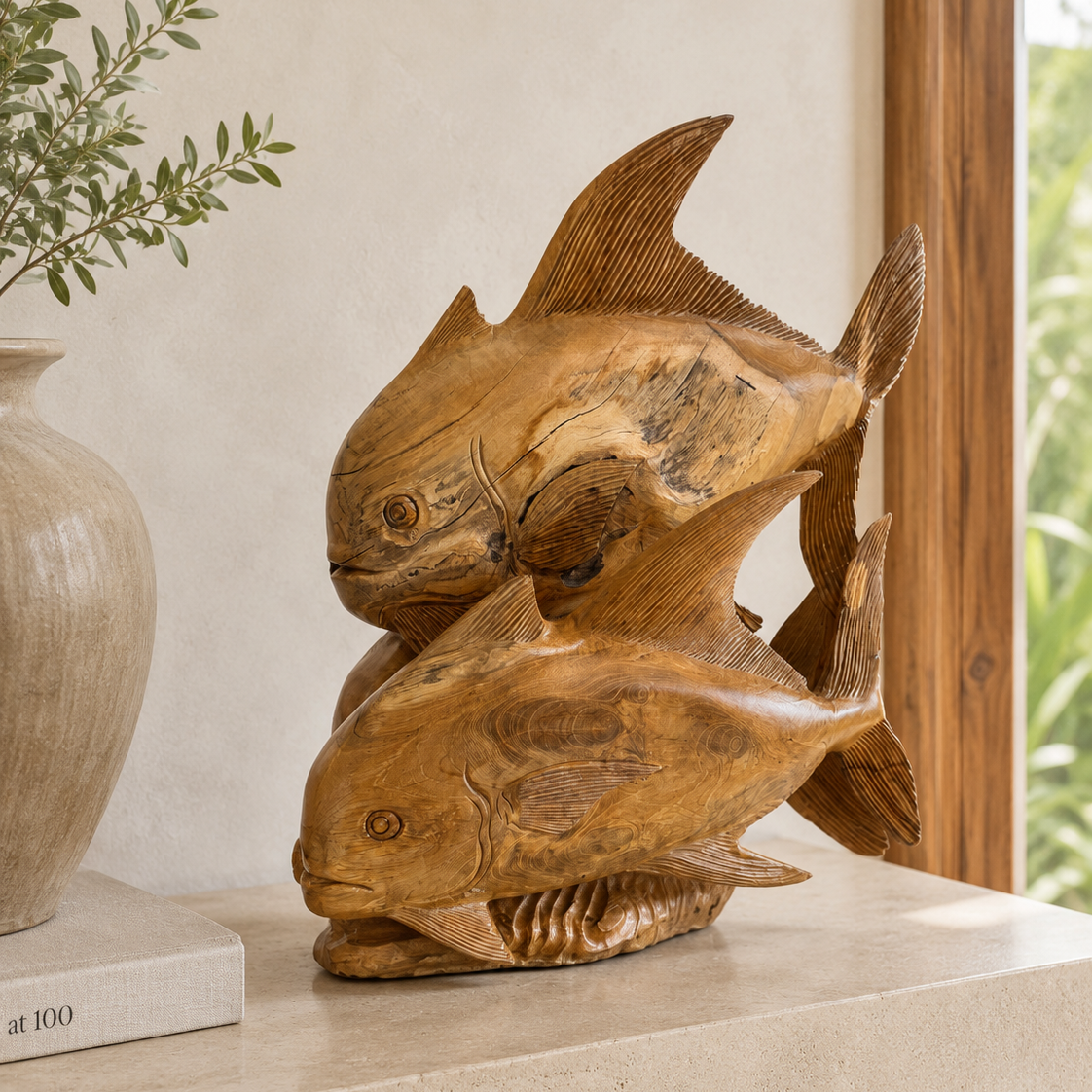 A carved wooden sculpture of two fish in a natural warm brown tone, displayed on a light stone shelf next to a ceramic vase and book.