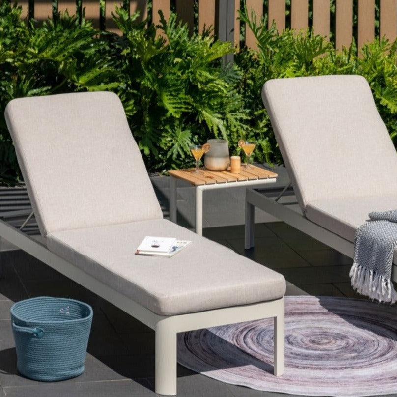 Two lounge chairs with cushions on a patio with a small table and plants in the background.