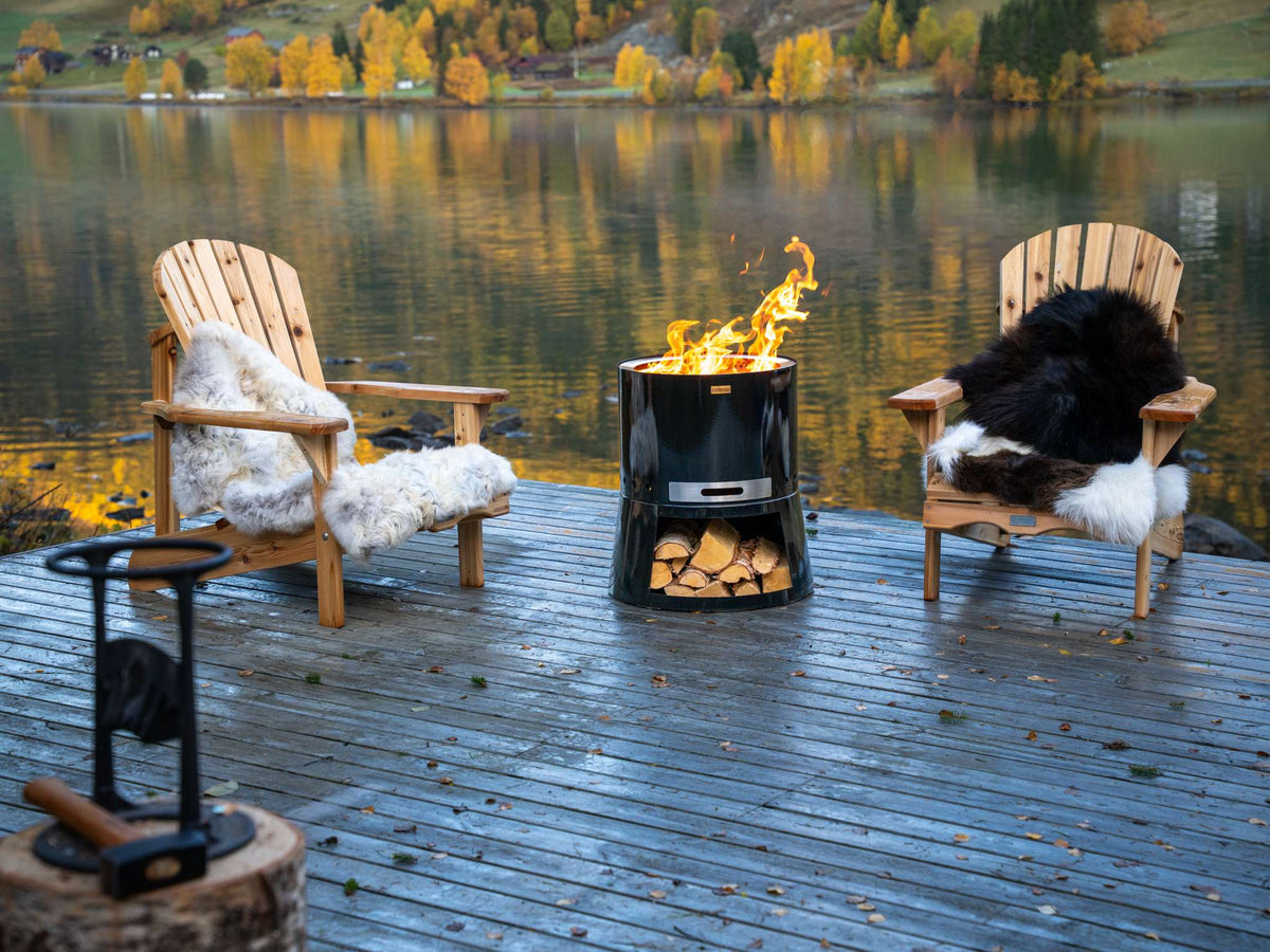 Two wooden chairs with fur cushions by a fire pit on a wooden deck by a lake.