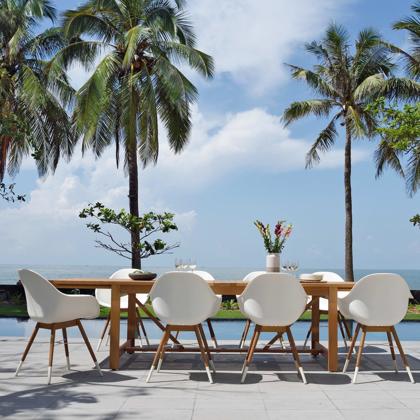 Outdoor dining setup with white chairs and a wooden table, surrounded by palm trees and a pool.