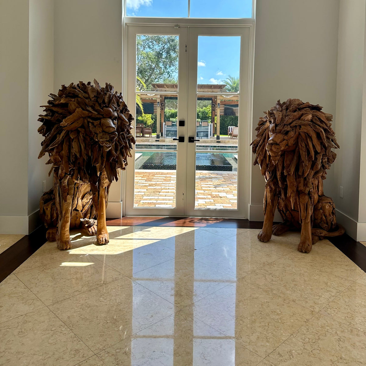 Two large sculptural lion statues made from natural teak, placed on either side of a glass double door in a bright entryway with tiled floors.