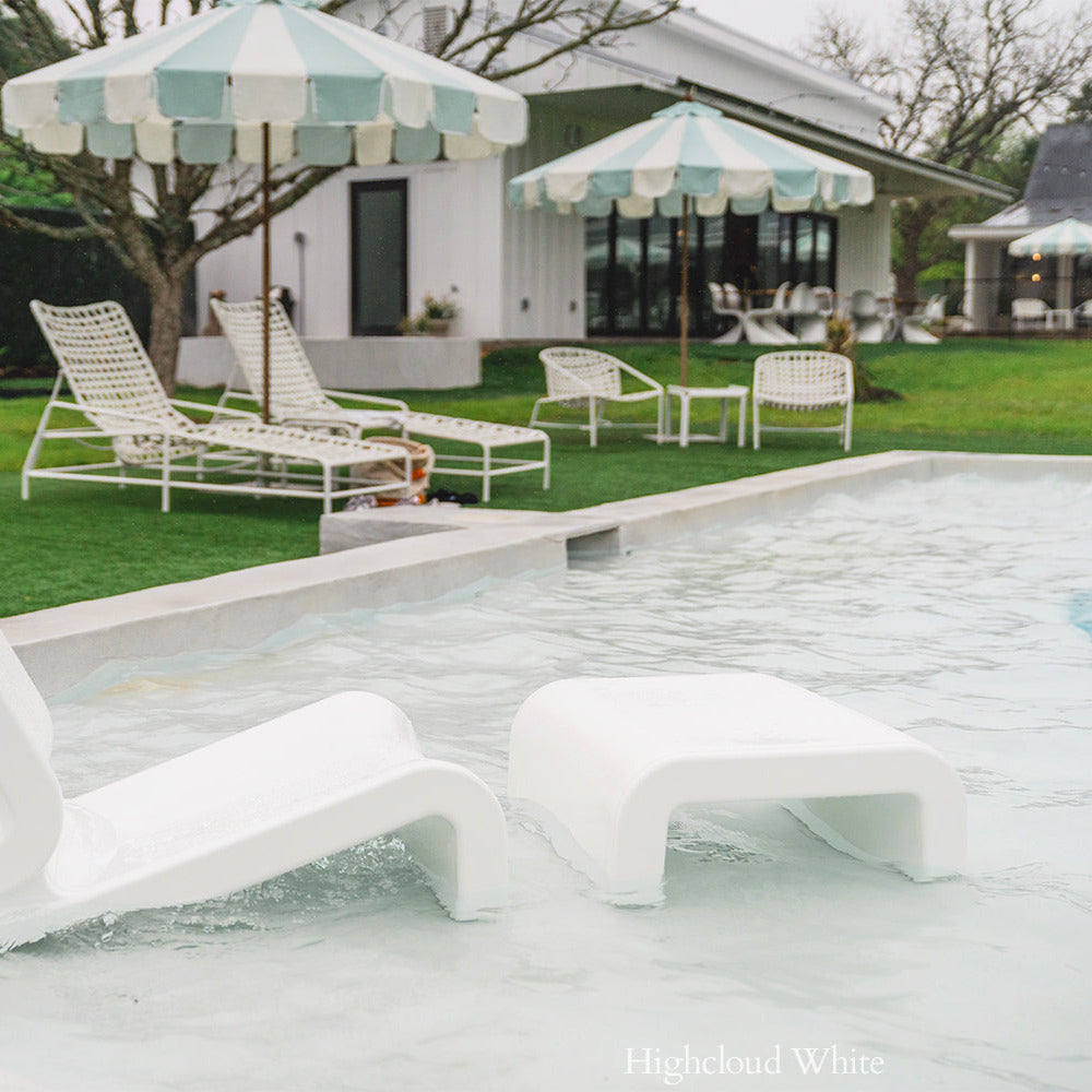 White lounge chairs by a pool with striped umbrellas and a house in the background.