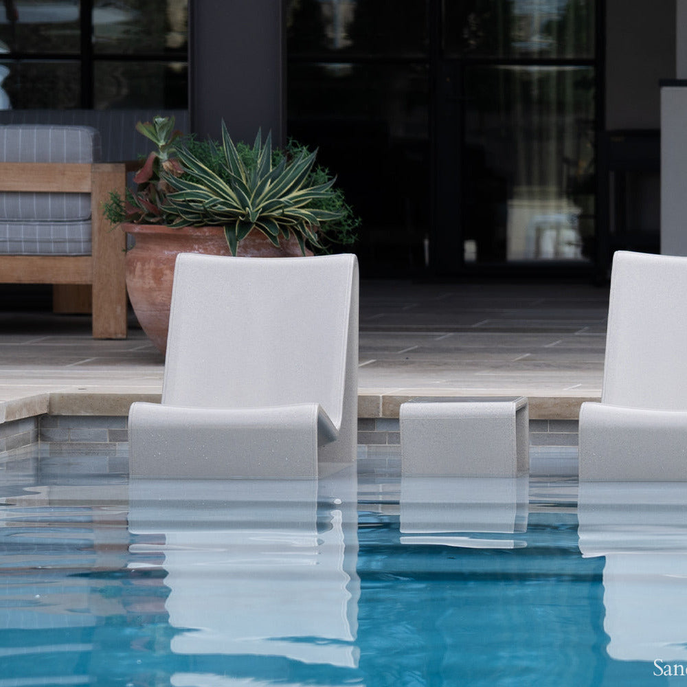 Two white chairs in a pool with a sandstone patio and potted plant in the background.
