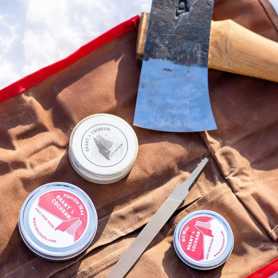 Three small round tins of Sharp & Coakman sharpening oil arranged on a brown leather tool roll with a wooden-handled axe, a metal file, and a metal rod, photographed outdoors on a snowy ground.