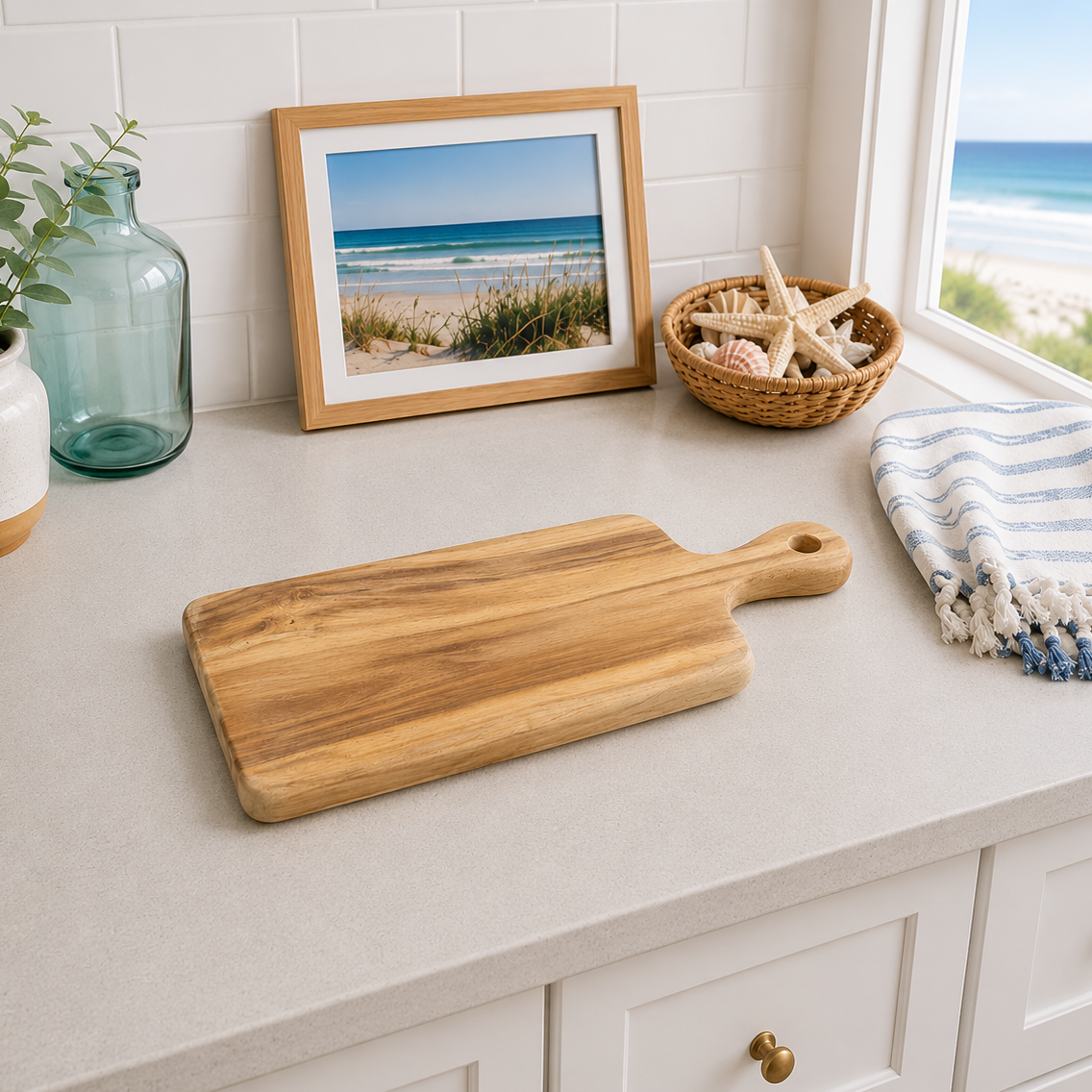 A rectangular wooden cutting board with a handle is displayed on a light gray countertop in a bright kitchen setting. The scene includes a blue-green glass bottle, a framed picture of a beach, a small basket with starfish, and a blue-and-white striped towel.