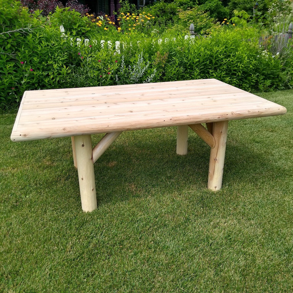A light natural wood dining table with thick cylindrical legs and a simple rectangular top, photographed outdoors on a green lawn with garden foliage and a white picket fence in the background.
